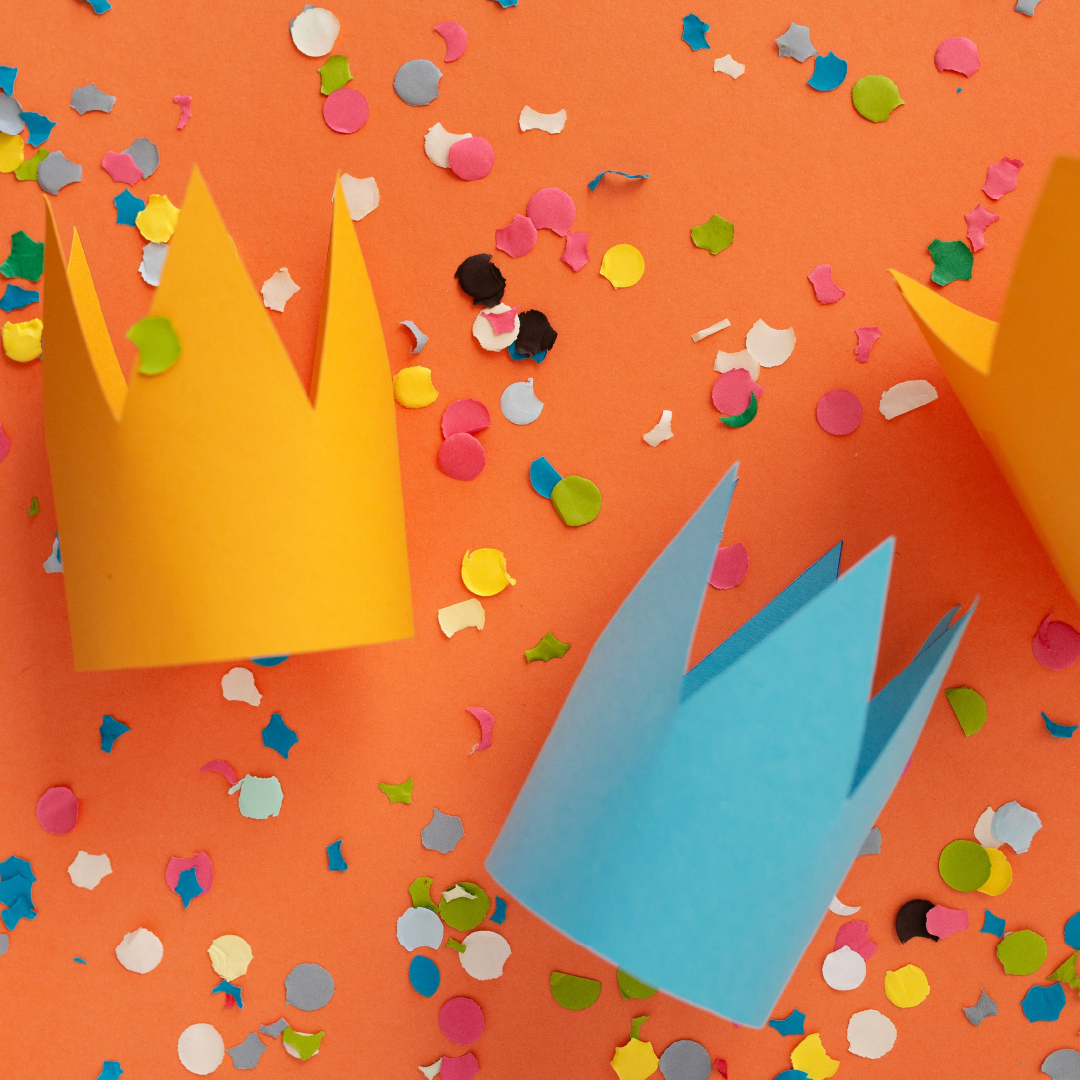 Yellow and blue paper crowns laid on an orange tablecloth covered in colourful confetti.
