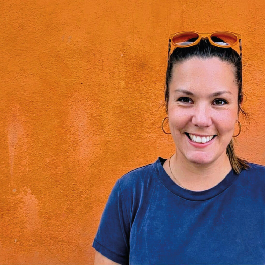 Becky (from Becky Whiting Communications) headshot wearing a blue t-shirt against an orange wall