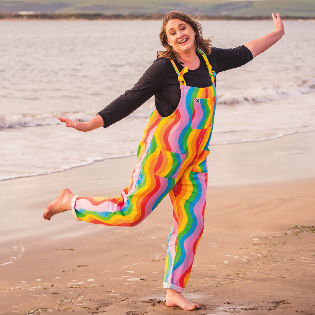Siobhan Strode (from This Sister Speaks) playfully dancing on a beach, wearing rainbow-coloured dungarees.